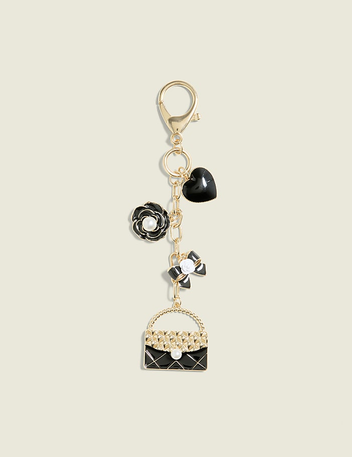 A gold keychain with charms of a black quilted handbag, a white and black bow, a black rose with a pearl, and a black heart hangs against a plain cream background.