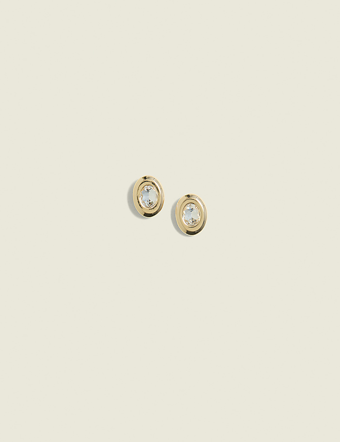 A pair of gold oval earrings with clear stones are centered against a soft, pale yellow background.