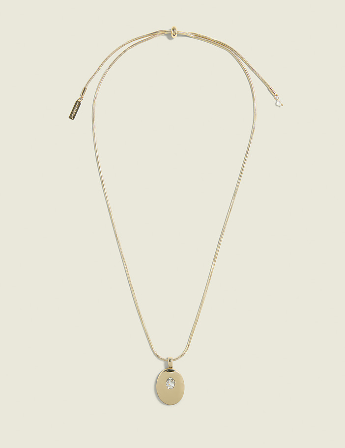 A delicate gold necklace with a circular pendant featuring a small inset gem hangs against a plain, light beige background.