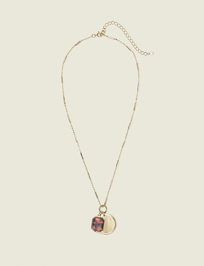 A gold necklace with a tortoise shell charm and a circular pendant hangs against a plain, light beige background.