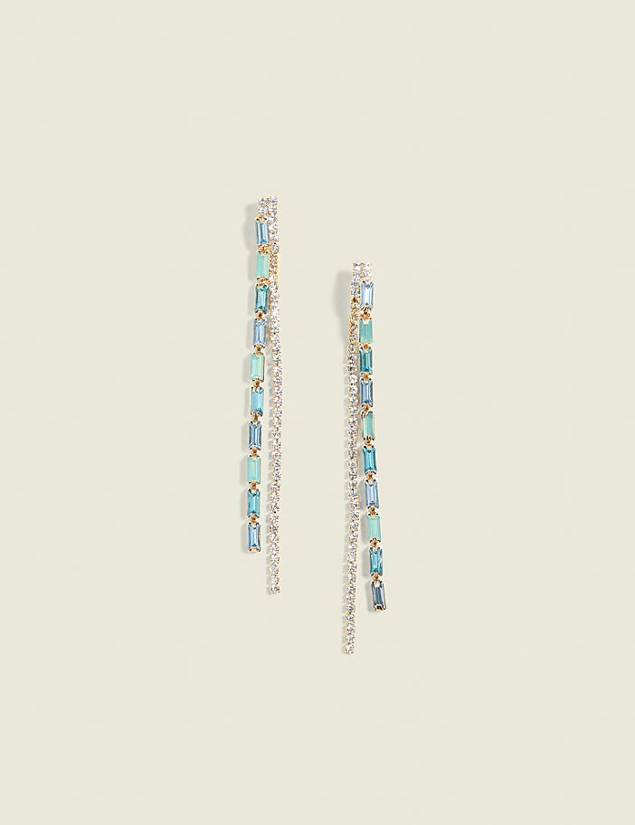 Two delicate gold drop earrings with alternating clear and blue gemstones hang against a soft, pale yellow background.