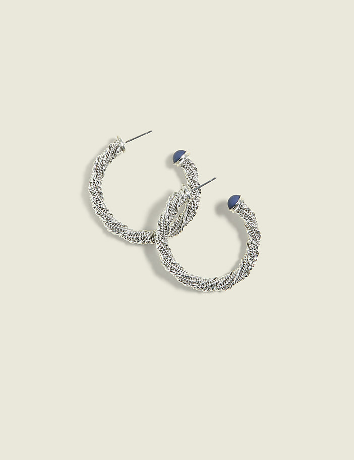 A pair of silver twisted hoop earrings with small blue stones rests on a pale yellow background.