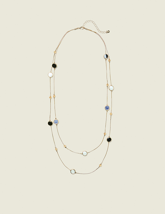 A delicate gold necklace with alternating black, white, and blue circular pendants and small amber beads is laid out against a plain, light beige background.