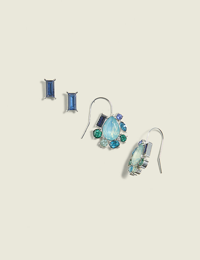 Two pairs of blue gemstone earrings, one set of studs and one set of dangling earrings, rest on a pale, textured background.