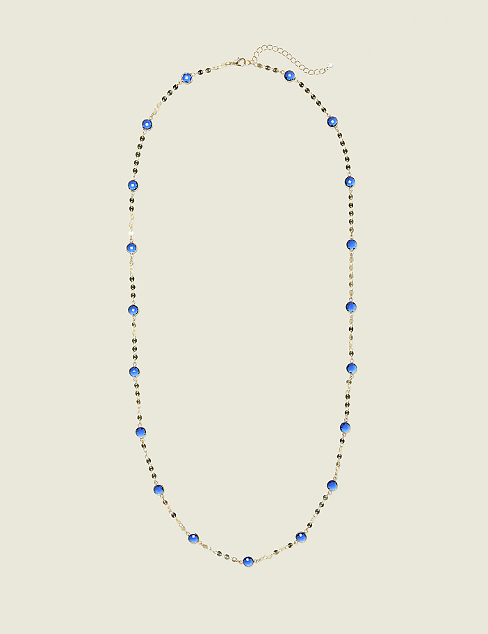 A delicate gold necklace with blue beads and small dark accents lies on a plain, light beige background.