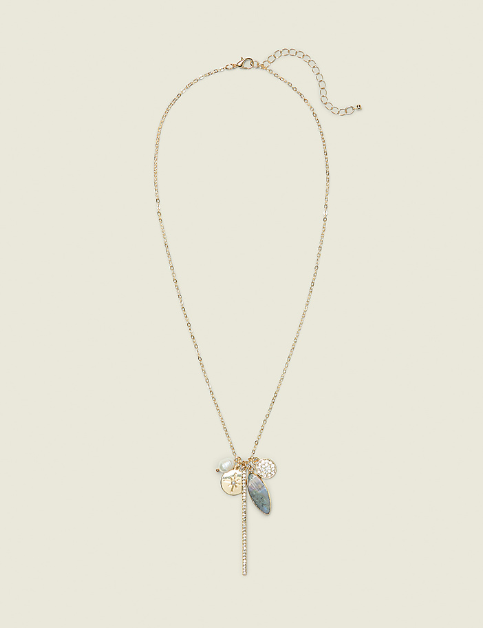 A delicate gold chain necklace with multiple charms, including a coin and a grey stone, hangs against a plain, light beige background.