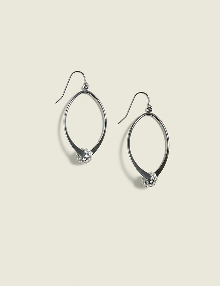 A pair of oval-shaped silver earrings with a small, sparkling ball at the bottom hang against a plain, light beige background.