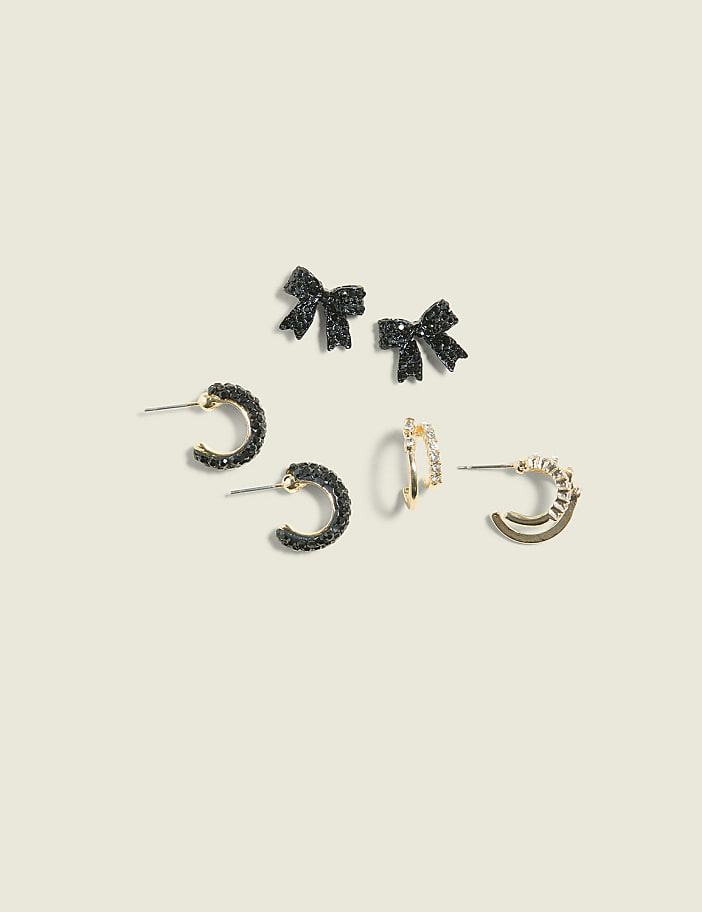 Several pairs of sparkling black and gold earrings, including bow studs and hoop earrings, are arranged on a pale cream background.