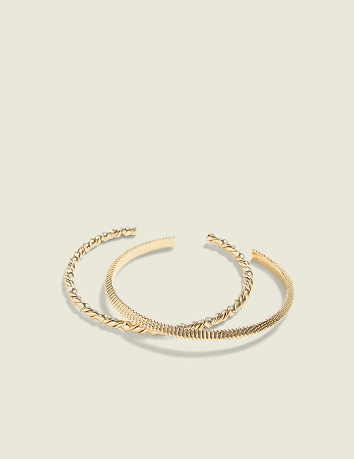 Two gold bangles, one with a twisted rope design and the other with a ribbed texture, rest on a plain, light-colored background.