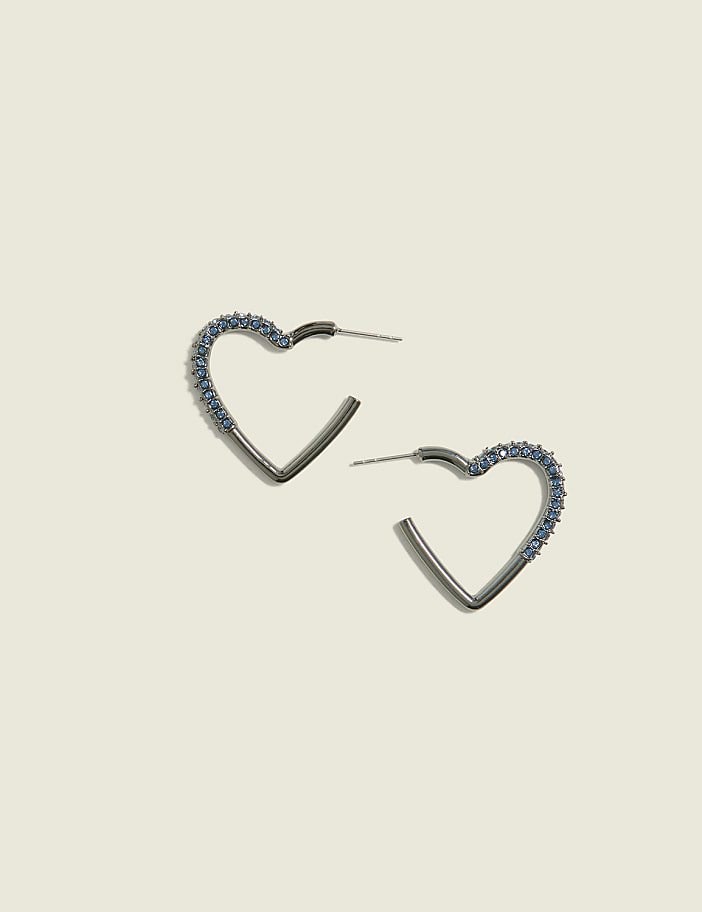 Two silver heart-shaped hoop earrings, one adorned with small blue gems, rest on a pale cream background.