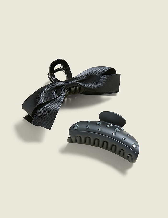 Two black hair clips, one with a satin bow and the other adorned with rhinestones, rest on a plain, light-colored background.