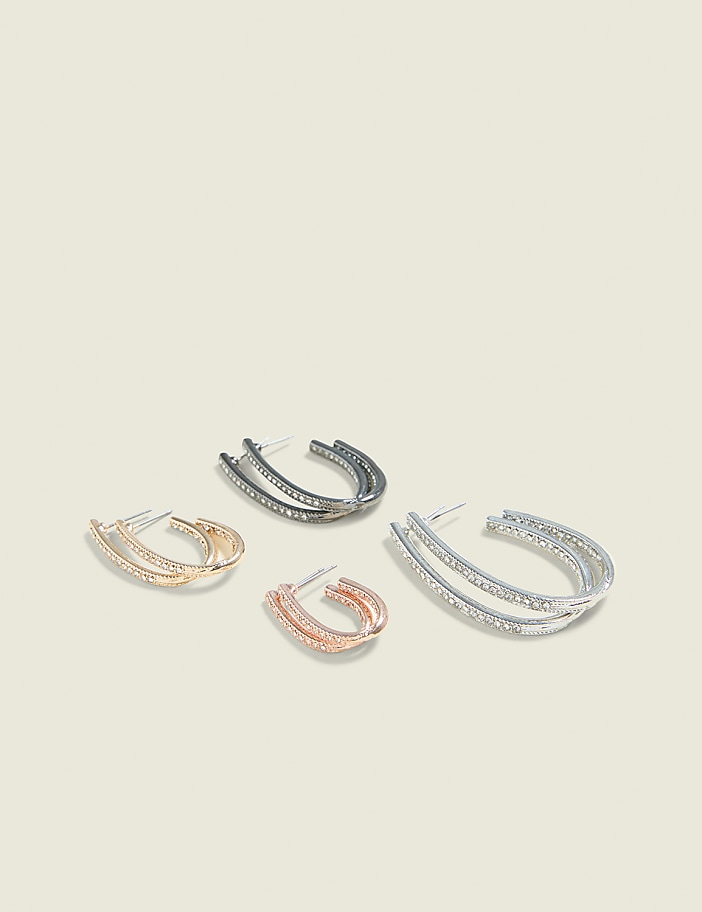 Four pairs of diamond-encrusted hoop earrings, in rose gold, silver, and gunmetal finishes, are arranged on a plain, light beige background.