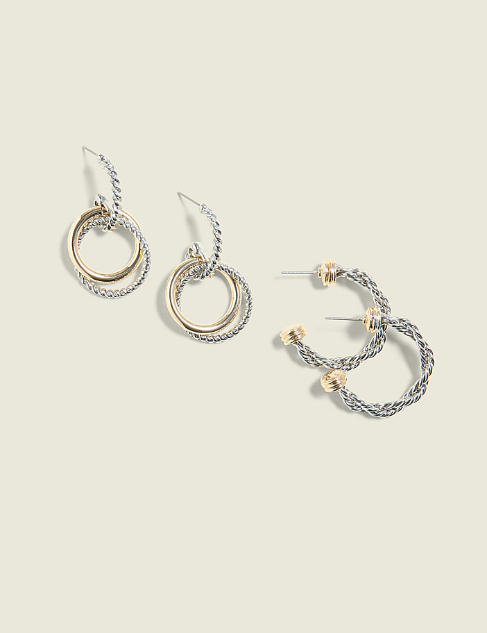 Two pairs of two-toned hoop earrings, one set dangling and the other lying flat, are displayed against a pale, textured background.