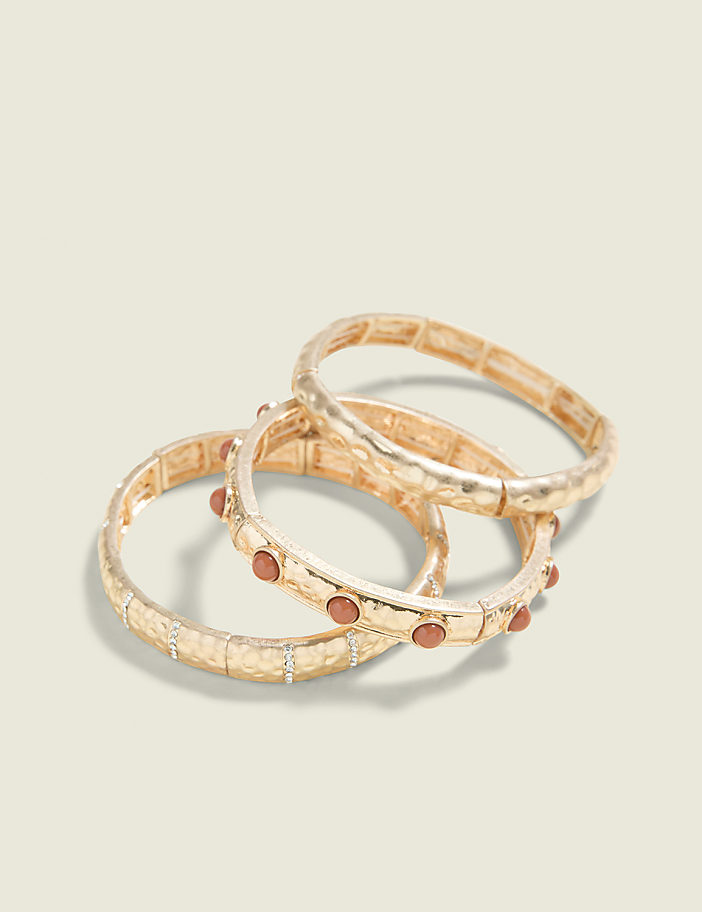 Three gold-toned bracelets, two adorned with small reddish-brown stones, are arranged on a plain, light beige background.