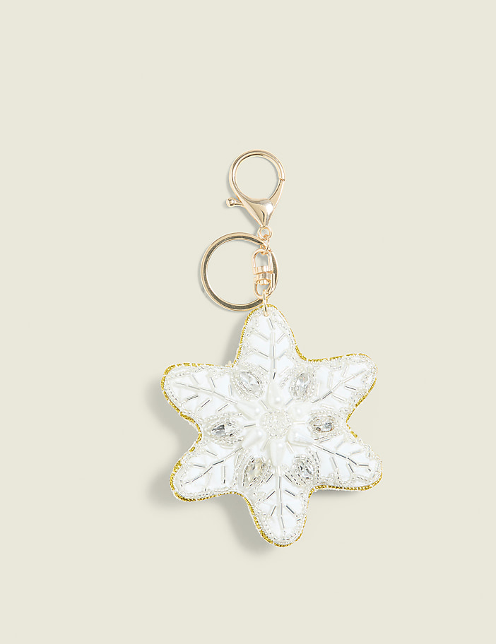 A white snowflake-shaped keychain with gold trim and rhinestones hangs against a plain cream background.