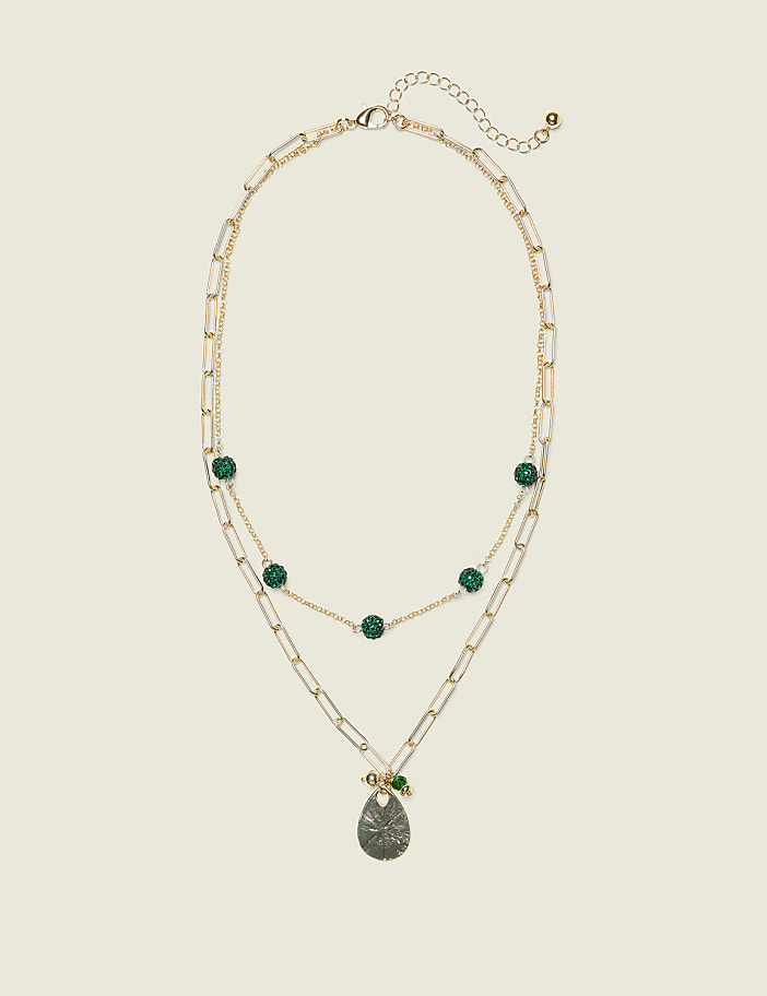 A layered gold necklace with green beads and a pendant hangs against a plain, light beige background.