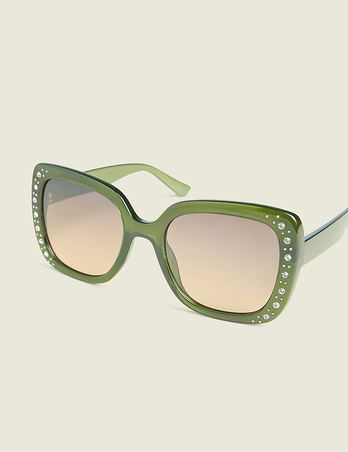 A pair of oversized, green, rhinestone-embellished sunglasses with gradient lenses rests against a plain, light-colored background.