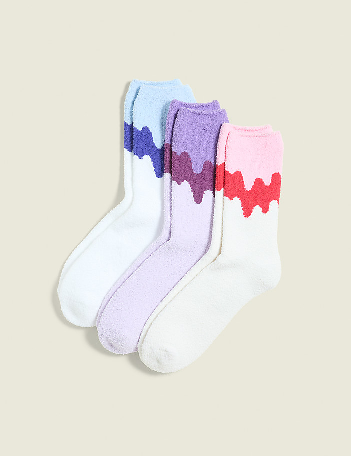 Three pairs of fluffy socks with colorful, dripping patterns are arranged in the foreground against a plain, light-colored background.