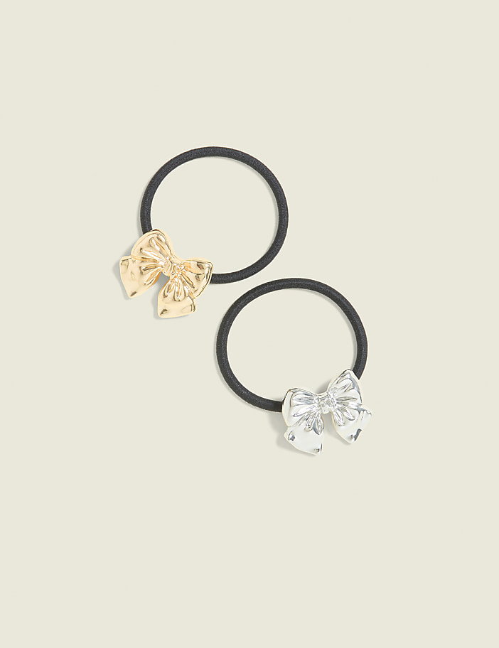 Two hair ties, one with a gold bow and another with a silver bow, rest on a plain, light-colored background.