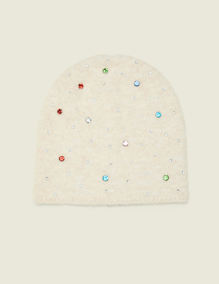 A fuzzy cream-colored beanie adorned with colorful rhinestones sits against a plain, light beige background.