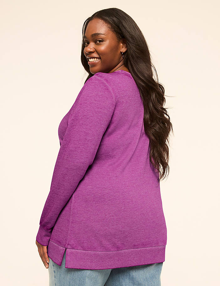 A smiling woman in a pink sweater and jeans stands against a plain, light-colored background.