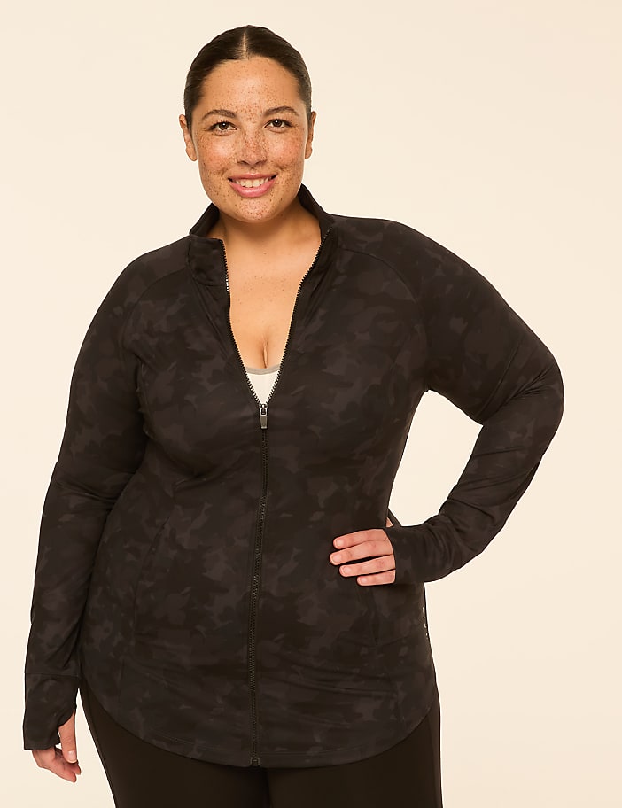 A smiling woman with freckles in a dark camouflage zip-up jacket stands against a plain, light-colored background.