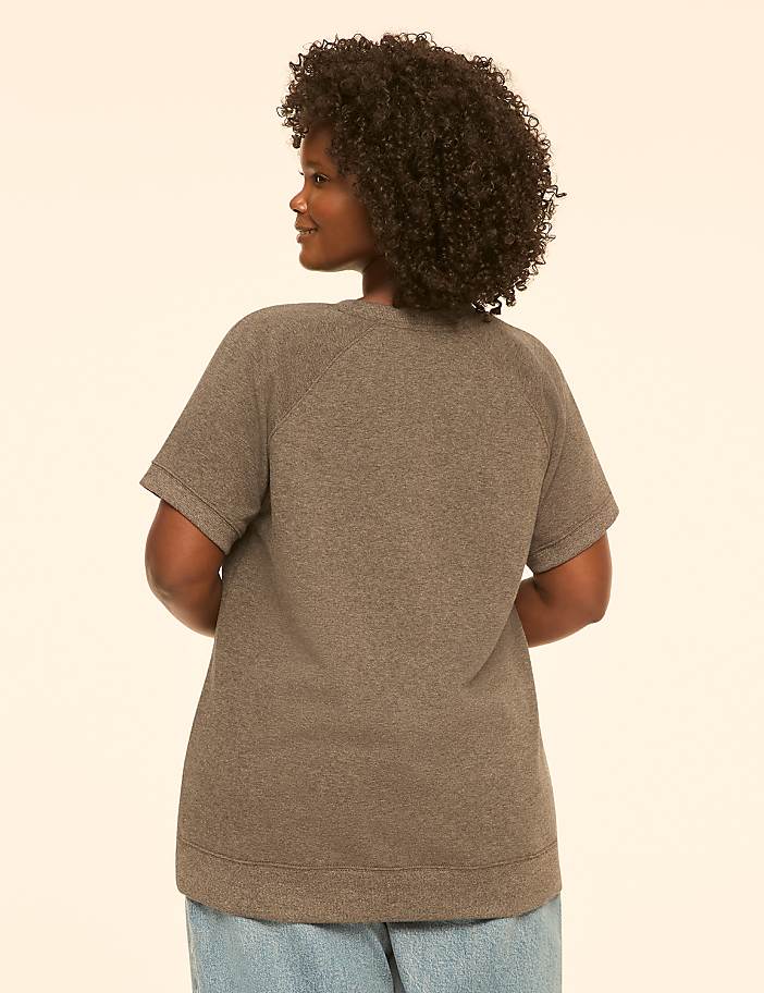 A woman with curly dark hair, wearing a gray t-shirt and blue jeans, stands against a plain cream background.