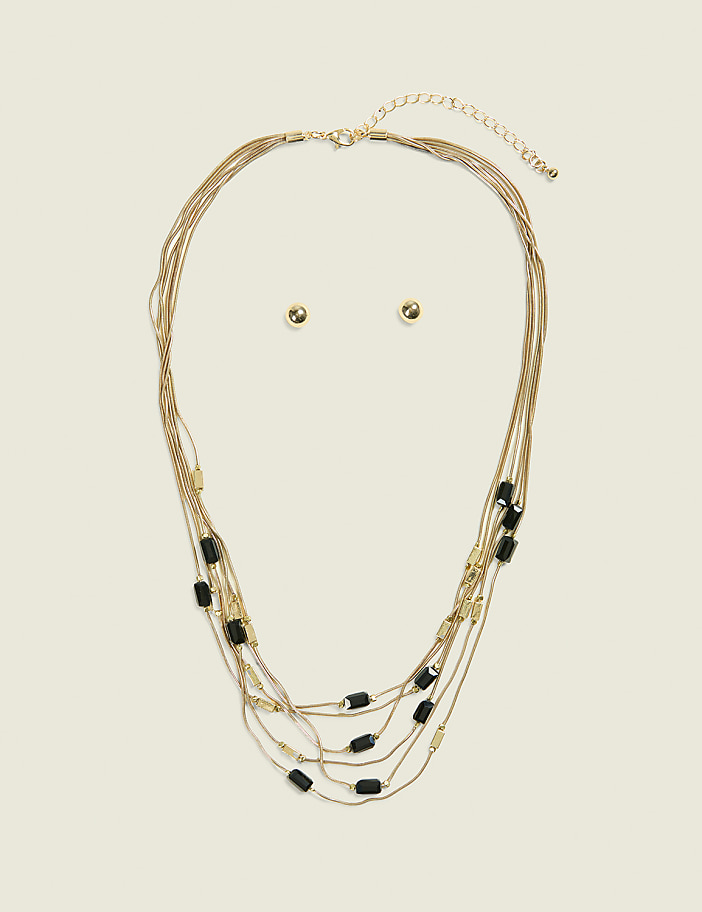A layered gold necklace with black beads and matching stud earrings are displayed against a plain, light beige background.
