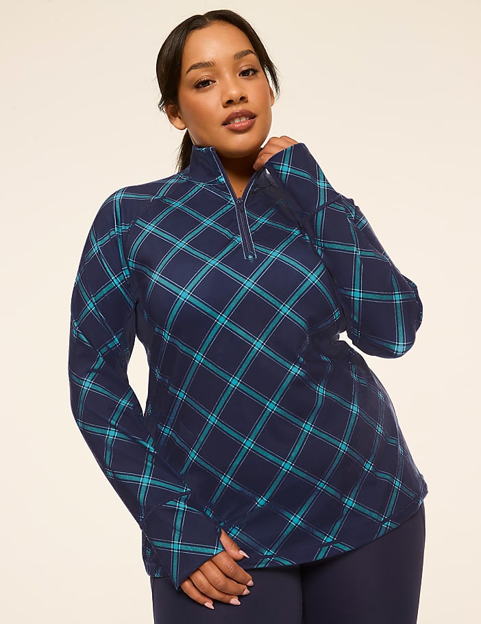 A woman in a blue plaid quarter-zip shirt and navy leggings poses against a plain, light beige background.