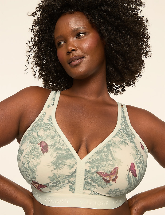A woman with dark, curly hair models a cream-colored bra with a delicate green and pink butterfly and foliage pattern against a plain, light background.