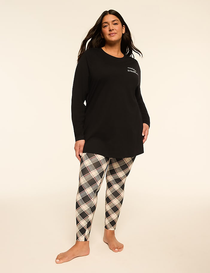 A woman stands in a black long-sleeved top and plaid leggings against a plain, light background.