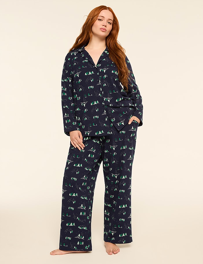 A woman with long red hair stands in a dark blue pajama set with a forest print, against a plain cream background.