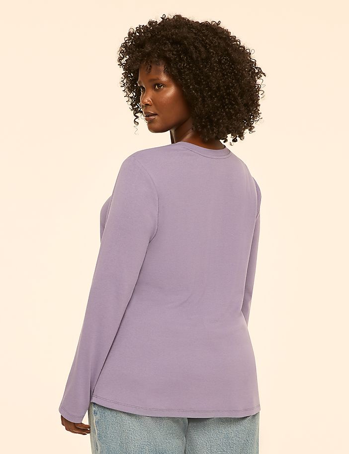 A woman with curly dark hair wearing a lavender long-sleeved shirt and jeans stands against a plain, light-colored background.