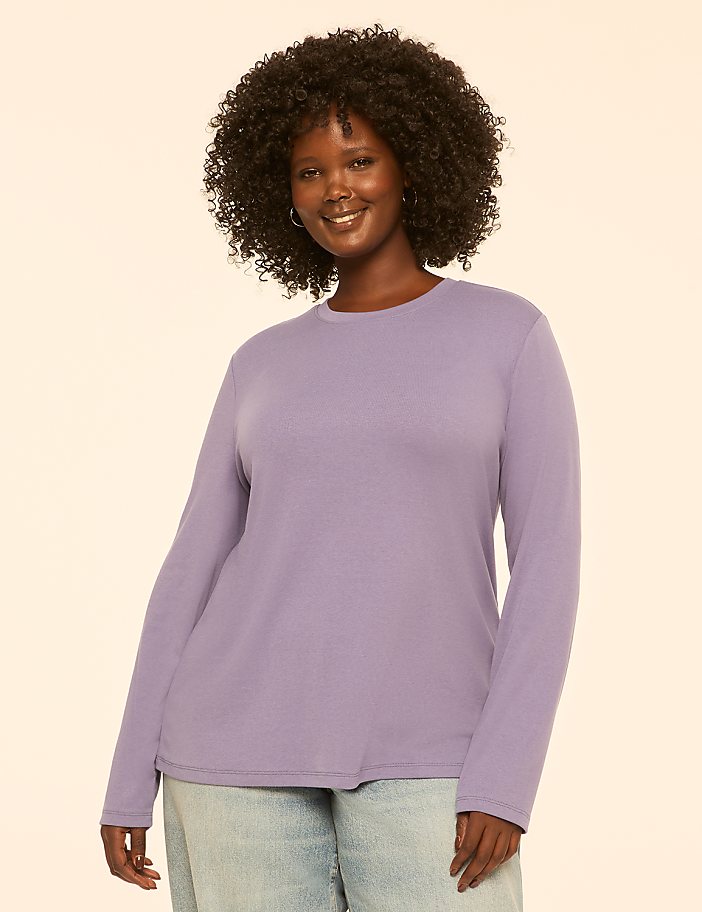 A smiling woman with curly dark hair wears a lavender long-sleeved shirt and light blue jeans against a plain, light-colored background.