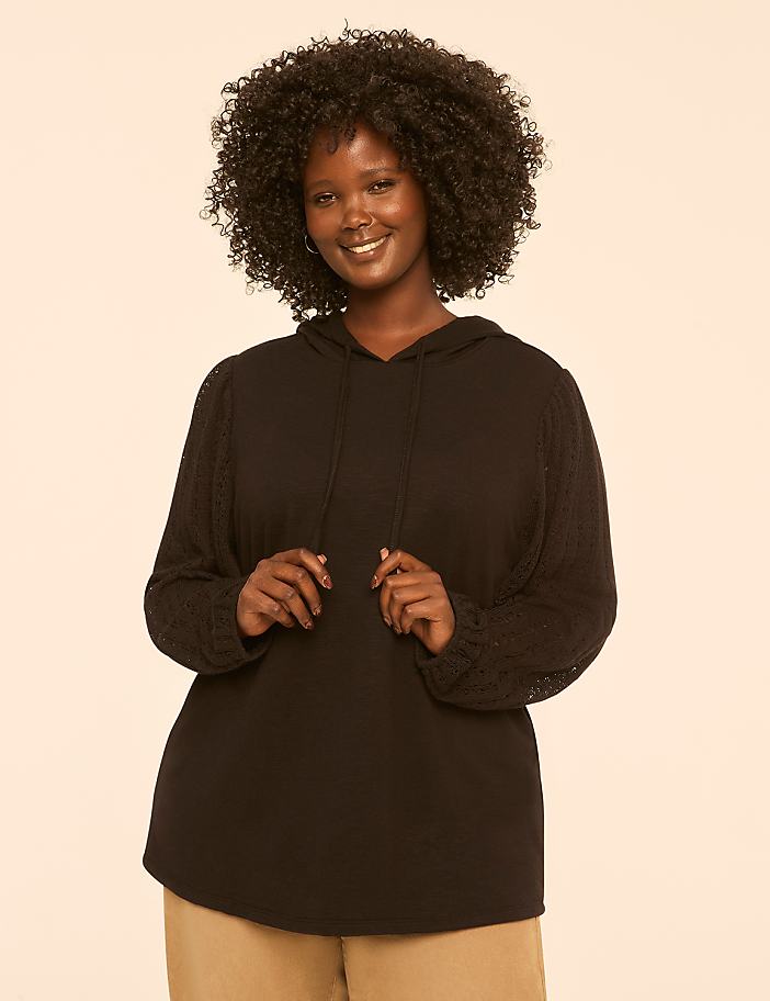 A smiling woman with dark, curly hair stands in the foreground wearing a black hooded sweatshirt, against a plain, light-colored background.