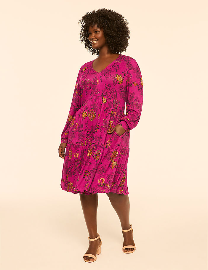 A woman in a vibrant pink floral dress stands against a soft, peachy background.