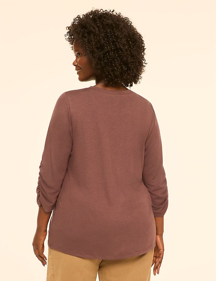 A woman with curly dark hair, wearing a ruched-sleeve mauve top and tan pants, stands against a plain cream background.