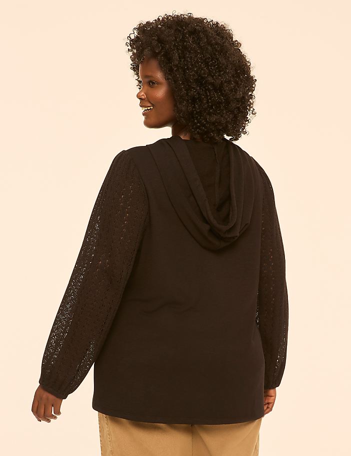 A woman with curly hair wears a black hooded sweatshirt with lace sleeves, standing against a plain, light-colored background.