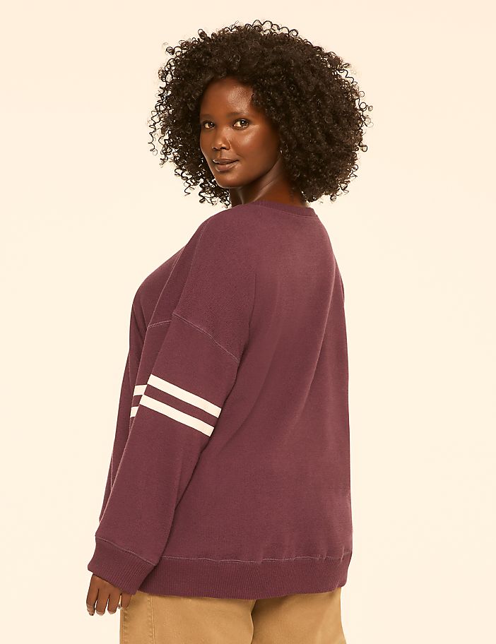 A woman with dark, curly hair poses in a maroon sweatshirt with white stripes on the sleeve, against a plain, light-colored background.