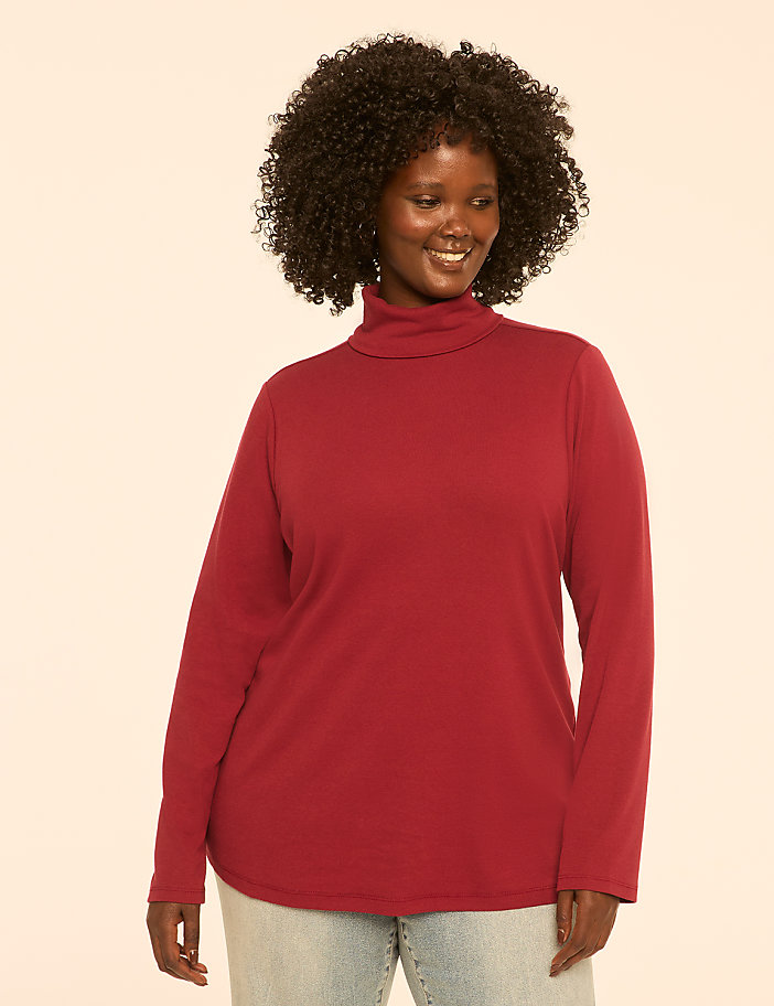 A smiling woman with curly dark hair wears a maroon turtleneck and light blue jeans against a plain cream background.