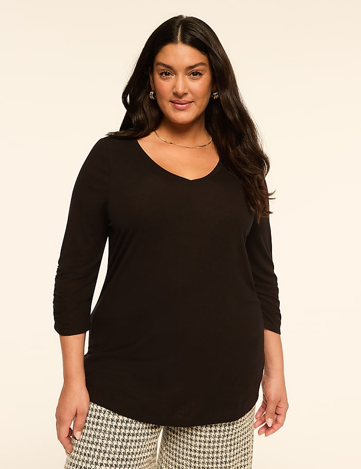 A woman in a black v-neck shirt and patterned pants stands against a plain, light-colored background.