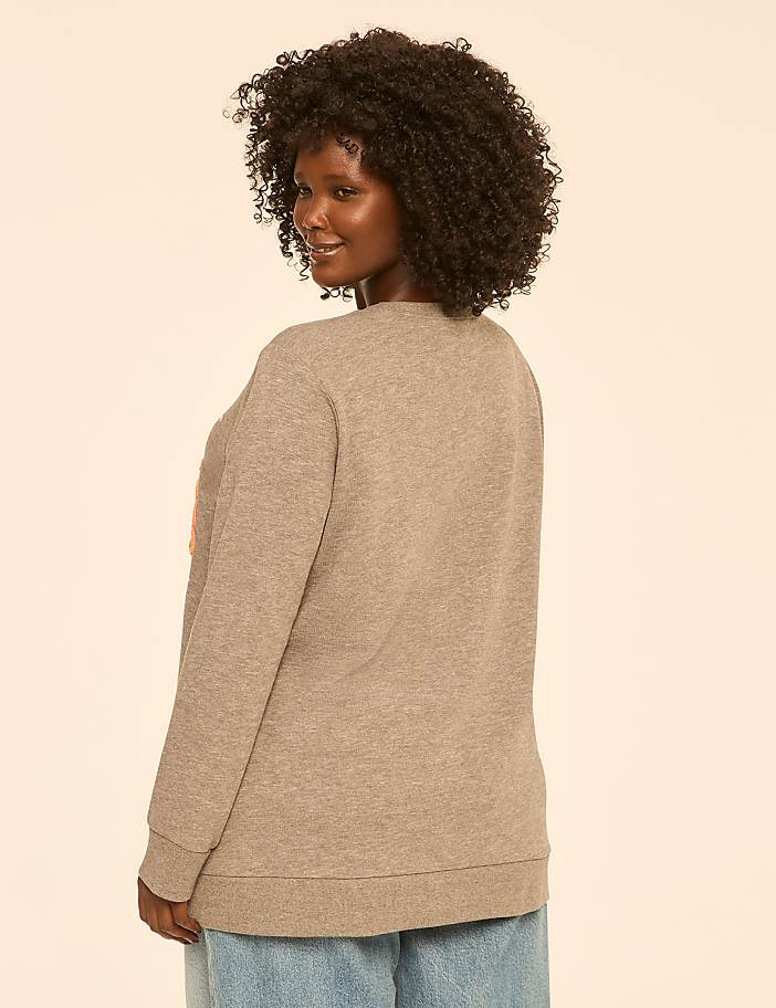 A woman with curly dark hair wearing a gray sweatshirt and blue jeans stands against a plain, light-colored background.