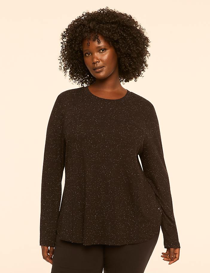 A woman with dark, curly hair and a black, sparkly long-sleeved shirt stands against a plain, light-colored background.