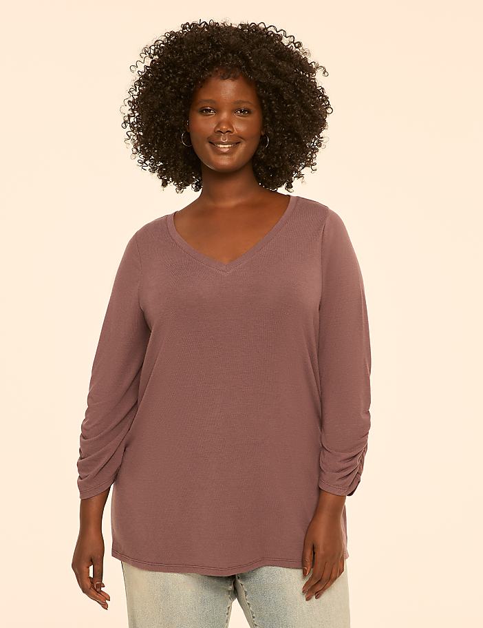 A smiling woman with curly dark hair and a mauve v-neck shirt stands against a plain, light-colored background.