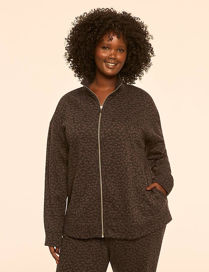 A smiling woman with curly dark hair stands in the foreground wearing a leopard-print zip-up jacket and matching pants against a plain, light-colored background.