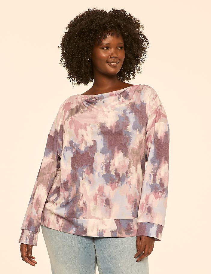 A woman with dark curly hair and a tie-dye sweatshirt stands against a plain, light-colored background.