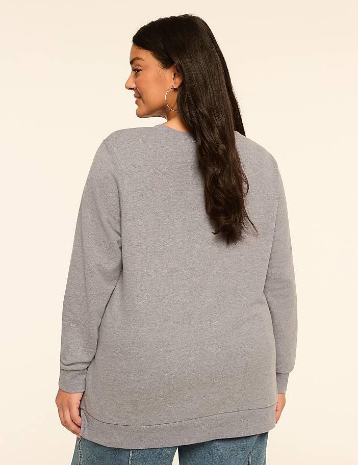 A woman in a gray sweatshirt stands against a plain, light-colored background.
