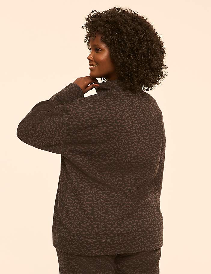A woman with curly dark hair, wearing a patterned dark top and pants, is shown from the back with her hand on her neck against a plain, light-colored background.