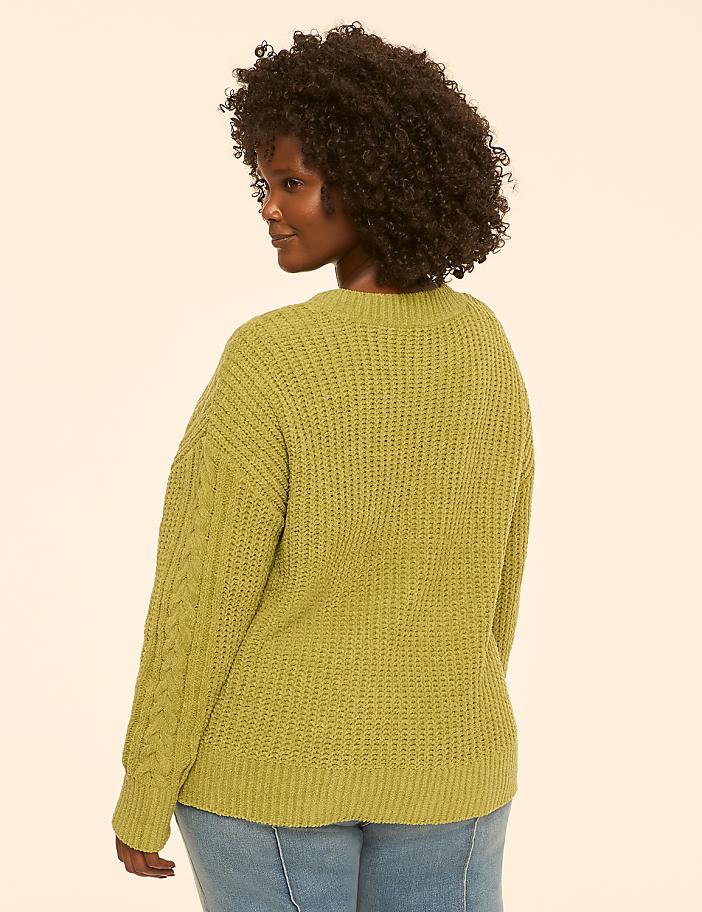 A woman in a green cable-knit sweater and jeans stands against a plain, light-colored background.