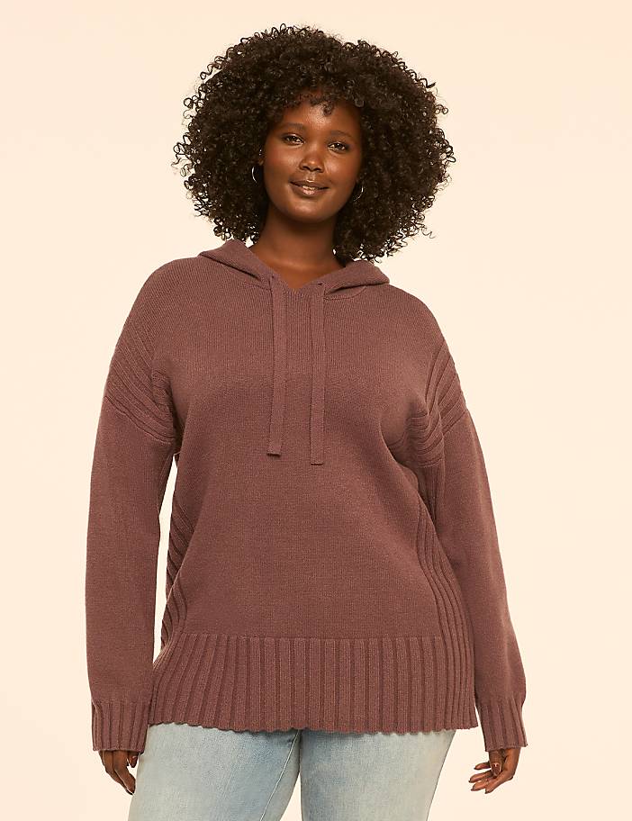 A woman with dark, curly hair smiles at the camera, wearing a mauve hooded sweater and light blue jeans against a plain, light peach background.